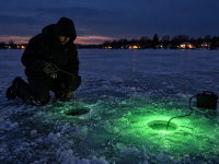Angler using a green underwater ice fishing light beside a fishing hole on a frozen lake